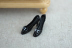 1:12 scale miniature women's high heel pumps in glossy black with small bows, one pair, shown on a gray fabric floor