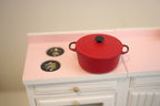 1:12 scale miniature large metal Dutch oven pot with lid in red with a black knob, shown on a pink and white stove