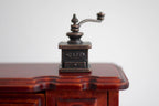 1:12 scale miniature metal coffee grinder in antique bronze with crank handle, shown on a mahogany wood table
