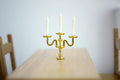 1:12 scale miniature gold metal candelabra with 3 arms and white candles, shown on a wood dining table