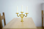 1:12 scale miniature gold metal candelabra with 3 arms and white candles, shown on a wood dining table