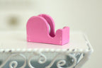 1:12 scale miniature tape dispenser in pink with a roll of tape, shown on a white table with a scrolled metal edge