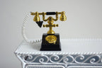 1:12 scale miniature metal rotary dial telephone in black and gold with white cord, shown on a white ornate table
