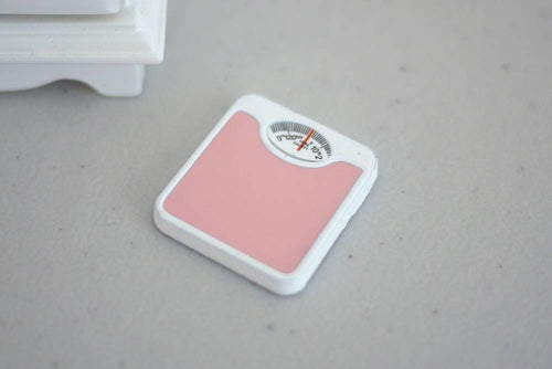 1:12 scale miniature bathroom scale in pink and white with a round dial, shown on a gray floor