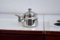 1:12 scale miniature metal kettle with lid in polished silver with curved spout, shown on a white stovetop