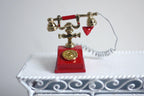 1:12 scale miniature metal rotary dial telephone in red and gold, ornate style with cord, on a white wire table