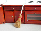1:12 scale miniature straw broom with white wood handle and red binding, shown leaning on a mahogany kitchen cabinet