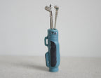 1:12 scale miniature golf bag in blue with black panel and 3 silver clubs, set of 4, standing on a gray surface