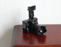 1:12 scale miniature retro camera in glossy black with flash unit and long lens, shown on a dark wood table