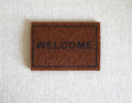 1:12 scale miniature light brown welcome doormat with black lettering and border, shown on a light linen surface