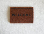1:12 scale miniature light brown welcome doormat with black lettering and border, shown on a light linen surface