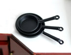 1:12 scale miniature frying pans in black, set of 3, large, medium and small, shown nested on a white counter