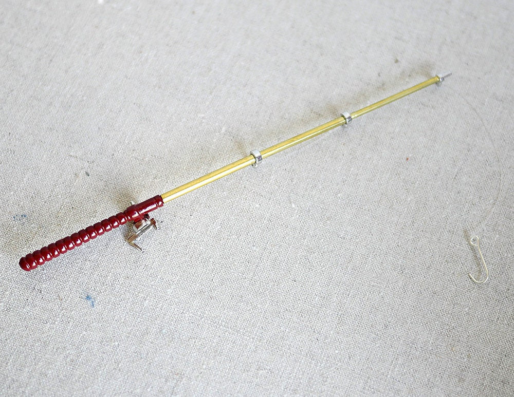 1:12 scale miniature metal fishing rod with reel, red handle and gold rod, shown lying on a white fabric surface