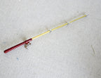 1:12 scale miniature metal fishing rod with reel, red handle and gold rod, shown lying on a white fabric surface