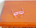1:12 scale miniature pink metal eyeglasses with clear lenses, shown folded open on a wood tabletop