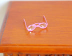 1:12 scale miniature pink metal eyeglasses with clear lenses, shown folded open on a wood tabletop