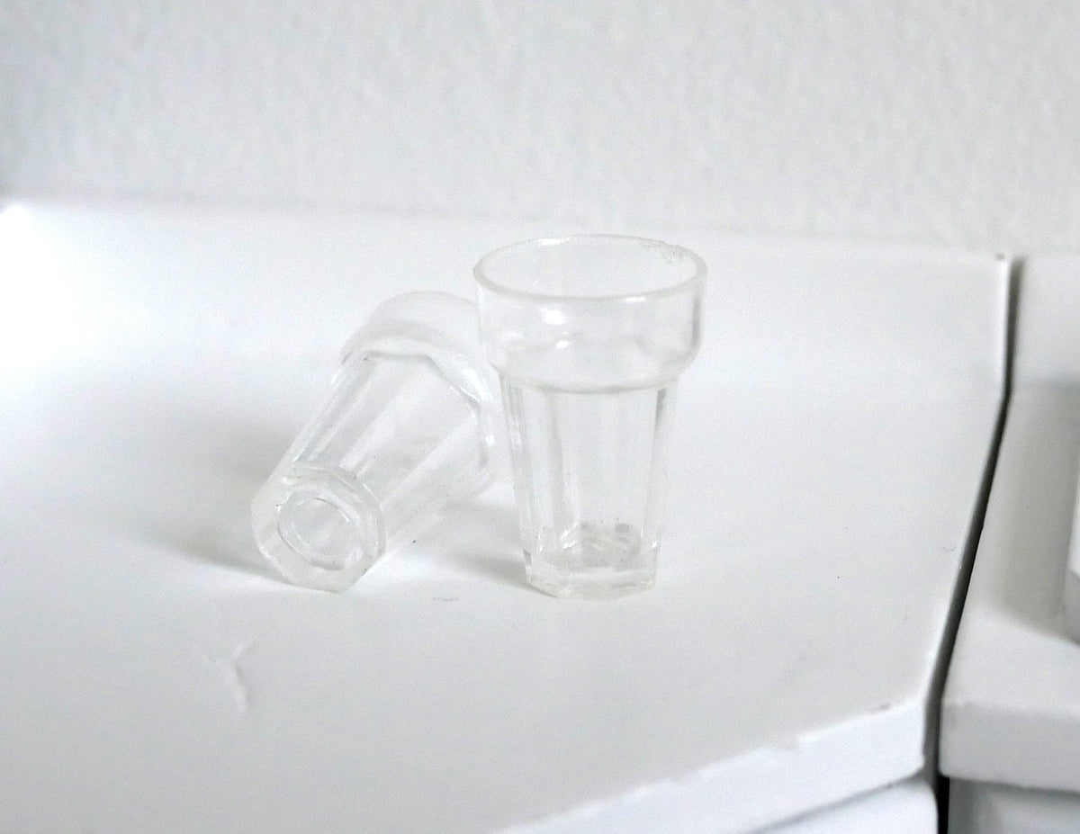 1:12 scale miniature clear tumbler glasses with faceted base, set of 2, one upright and one on its side on a counter
