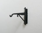 1:12 scale miniature metal hanging wall bracket with hook in black, scroll arm design, shown mounted on a white wall