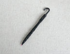 1:12 scale miniature folded umbrella in black with J hook handle, shown lying on a beige linen surface