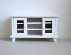 1:12 scale miniature wood TV stand in white with 2 glass pane doors and open center shelves, shown on a gray floor