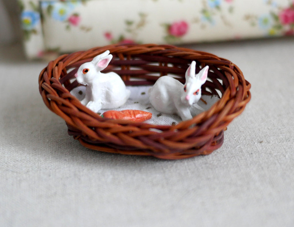 1:12 scale miniature white bunnies in a brown rattan bed with a carrot, two rabbits on a white cushion, on a beige carpet