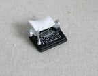 1:12 scale miniature black typewriter with silver keys and a white paper sheet, shown on a linen surface