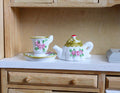 1:6 scale miniature porcelain teapot with cup and saucer, pink rose pattern with gold lid, shown on a wood hutch
