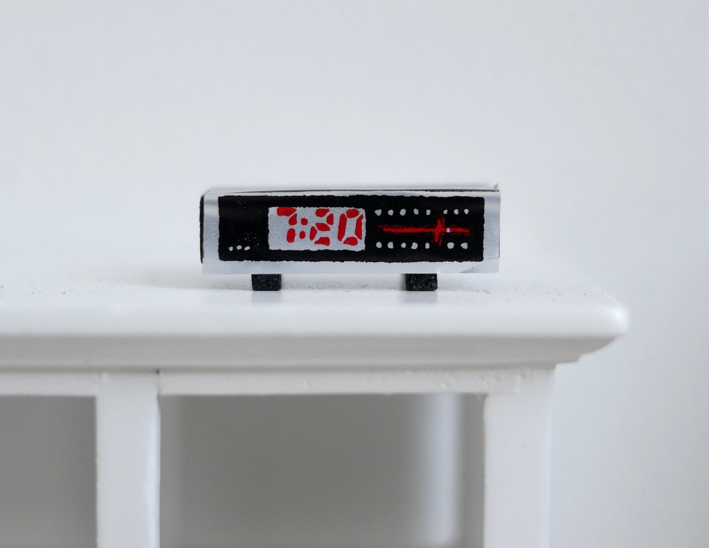 1:12 scale miniature digital alarm clock radio in black with red display digits and silver trim, on a white table