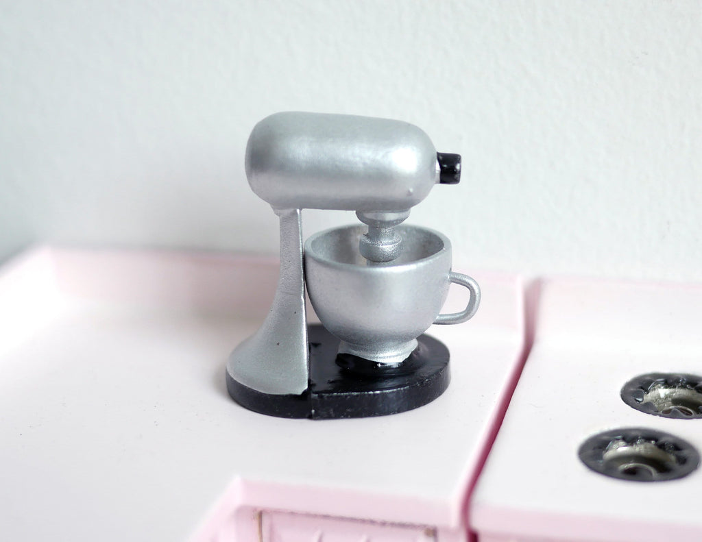 1:12 scale miniature stand mixer in silver with bowl on a black base, shown on a white and pink kitchen counter