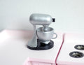 1:12 scale miniature stand mixer in silver with bowl on a black base, shown on a white and pink kitchen counter