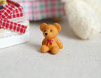 1:12 scale miniature super tiny teddy bear in brown with a red bow, sitting on the floor beside a bed