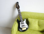1:12 scale miniature electric guitar in black and white with wood tone neck, shown leaning on a green sofa