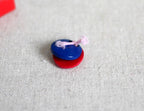 1:12 scale miniature finger castanets in blue and red joined with a pink cord, shown on beige linen fabric
