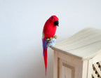 1:12 scale miniature red parrot figure with blue wing and tail feathers, shown perched on a wood cabinet