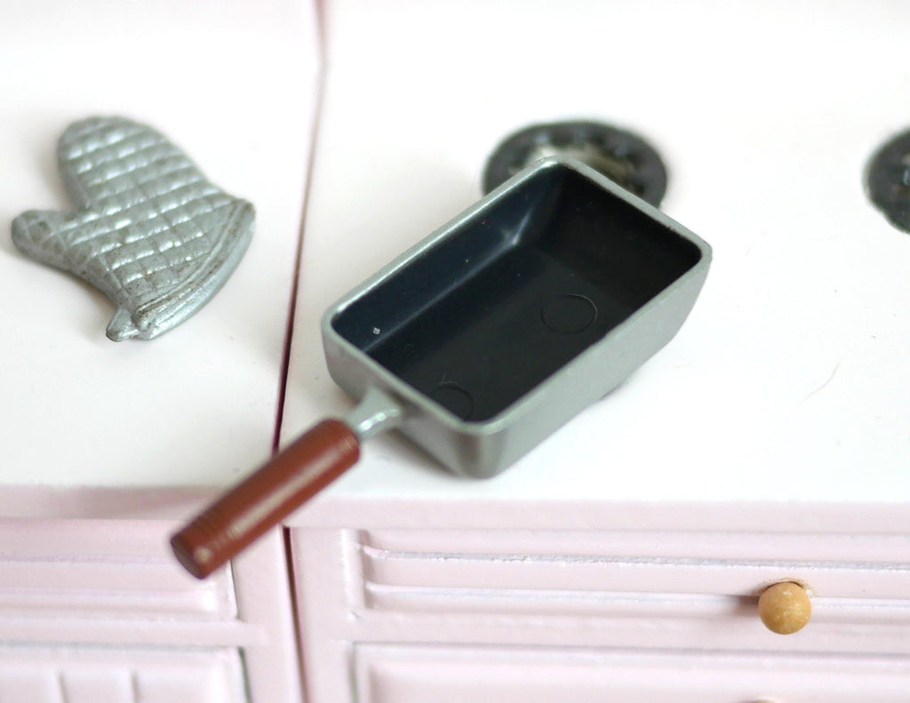 1:12 scale miniature black rectangular omelette pan with wood tone handle, shown on a pink kitchen stove