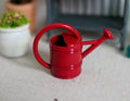 1:12 scale miniature red metal watering can with long spout and hoop handle, shown beside a potted plant
