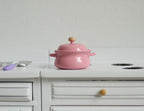 1:12 scale miniature pink metal cooking pot with lid, 2 side handles and a wood knob, shown on a white kitchen counter