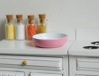 1:12 scale miniature ceramic shallow bowl in pink with a white inside, shown on a white kitchen counter by spice jars