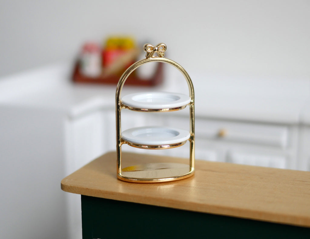1:12 scale miniature gold metal 2 tier serving stand with white plates and bow top handle, shown on a kitchen counter