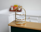 1:12 scale miniature gold metal 2 tier serving stand with white plates and bow top handle, shown on a kitchen counter