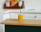 1:6 scale miniature cup of orange juice with clear lid and green straw, shown on a wood kitchen counter