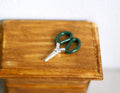 1:12 scale miniature scissors with green handles and silver blades, shown on a wood side table