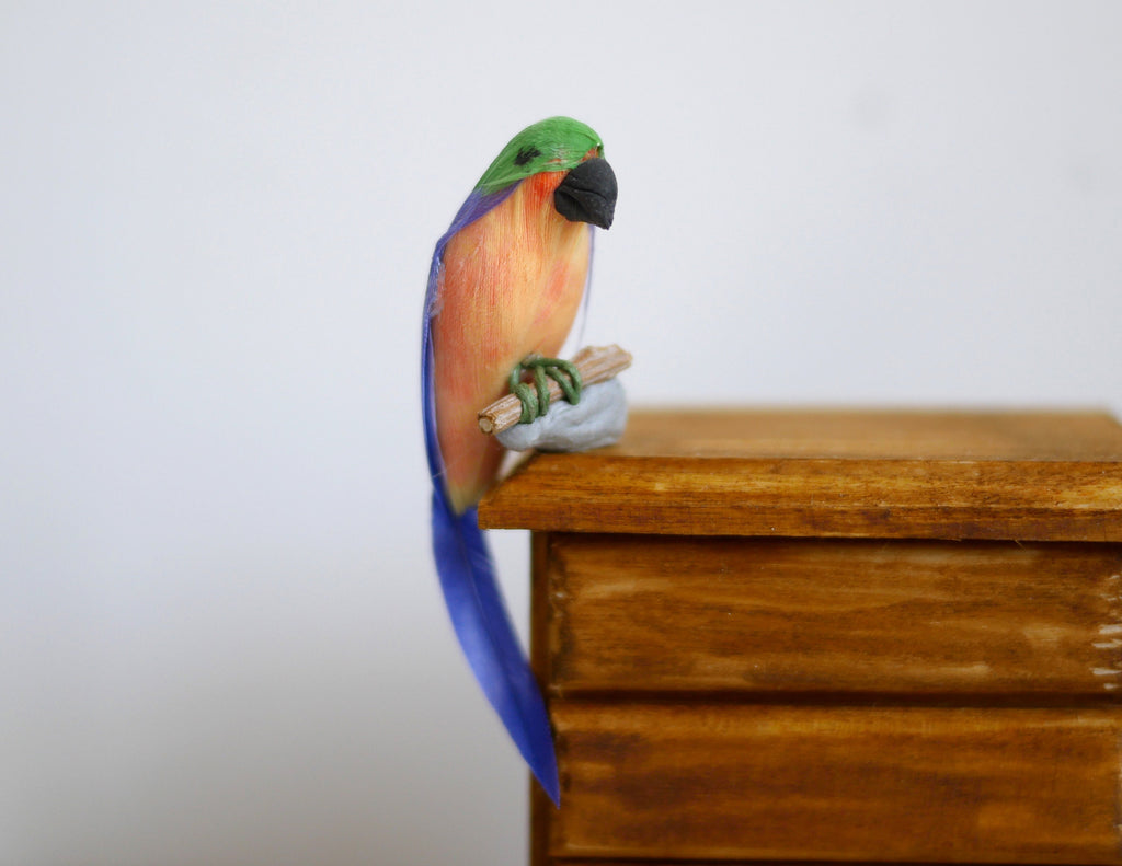 1:12 scale miniature parrot figure edge sitter with green head, orange body and long blue tail, on a wood dresser