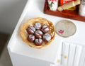 1:12 scale miniature wicker basket of glazed buns with powdered tops, shown on a white counter beside a coin