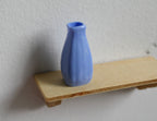 1:12 scale miniature ribbed vase in periwinkle blue with a narrow neck, shown on a small wood shelf