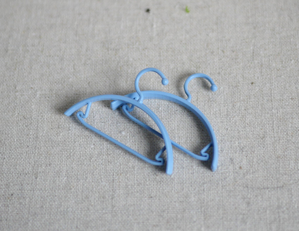 1:12 scale miniature clothes hangers in light blue, set of 2, shown lying on a beige fabric surface