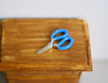 1:12 scale miniature stationery scissors with blue handles and silver blades, shown on a wood table