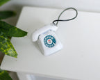1:12 scale miniature metal rotary dial telephone in white with teal dial and black cord, on a white table