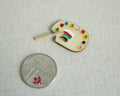 1:12 scale miniature oval artist palette with paint dabs and brush, shown on linen beside a coin for scale