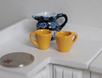 1:12 scale miniature coffee mugs with handles in yellow, set of 2, shown on a white counter beside a blue floral jug